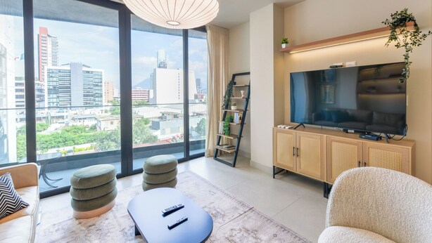 A modern living room with a large window, a flat-screen TV, a sofa, ottomans, and a coffee table. - Niza (Monterrey)
