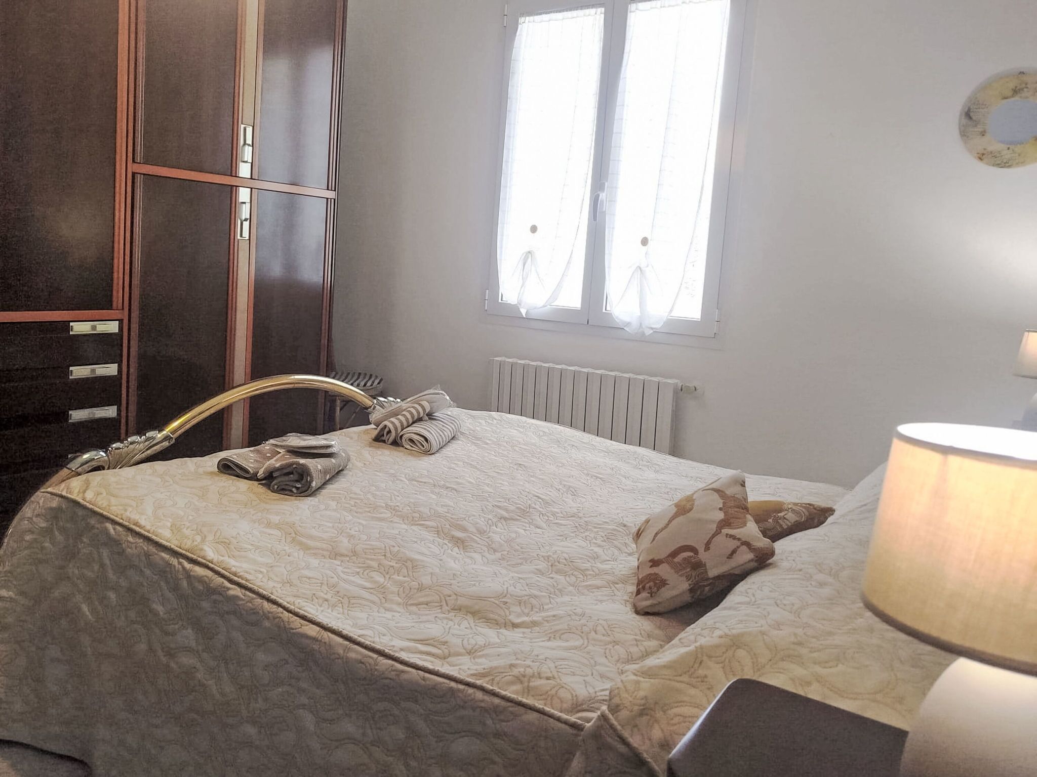 A bedroom with a bed, a radiator, a window with curtains, a wardrobe, and a lamp.