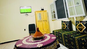A room with a television, a table with a patterned tablecloth, a sofa with patterned cushions, a wooden cabinet, and a window with blinds. - Amazigh host Taroudannt (Taroudant)