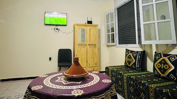 A room with a dining table, a television mounted on the wall, a window with blinds, and a patterned rug. - Amazigh host Taroudannt (Taroudant)