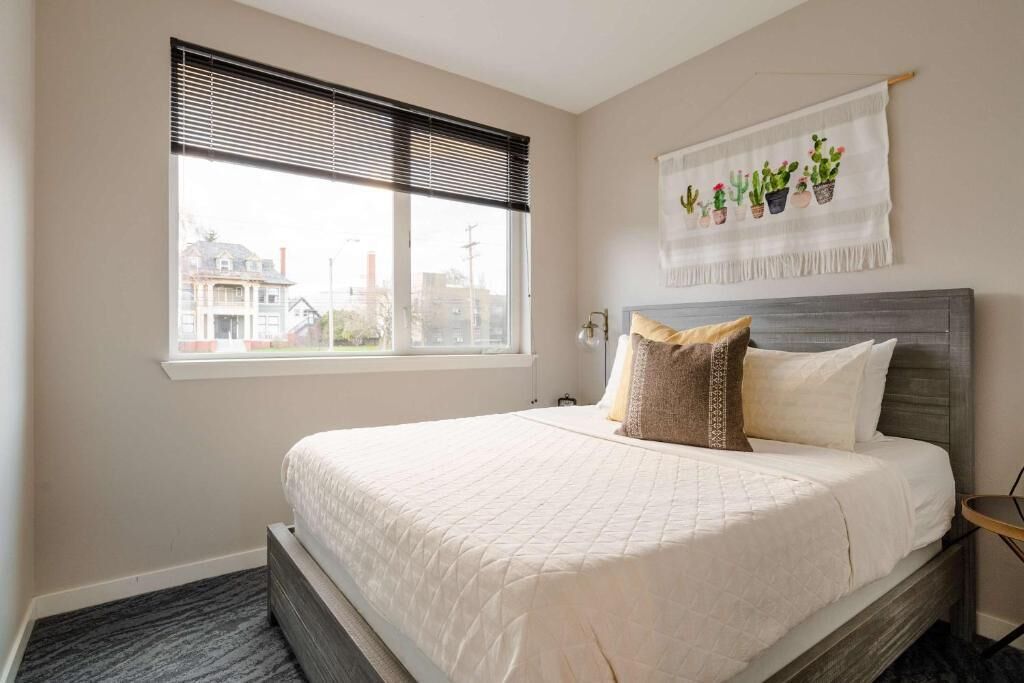 A bedroom with a bed, a window with blinds, a wall-mounted plant decoration, and a view of buildings outside.