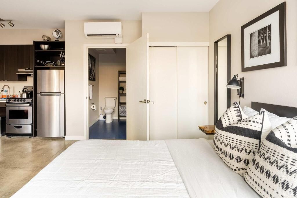 A bedroom with a bed, pillows, a nightstand, a wall-mounted lamp, and a door leading to a bathroom.
