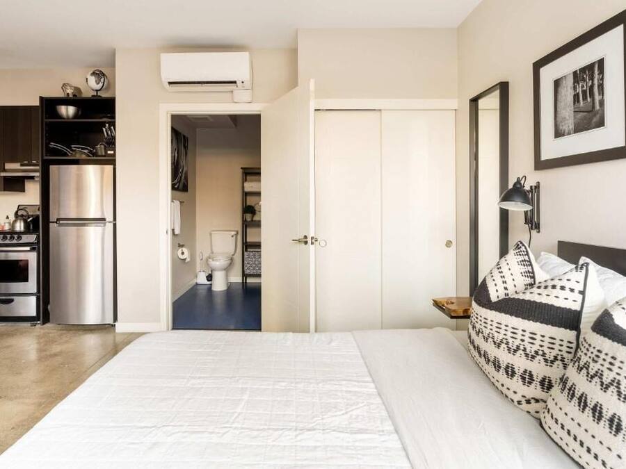 A bedroom with a bed, pillows, a nightstand, a wall-mounted lamp, and a door leading to a bathroom.