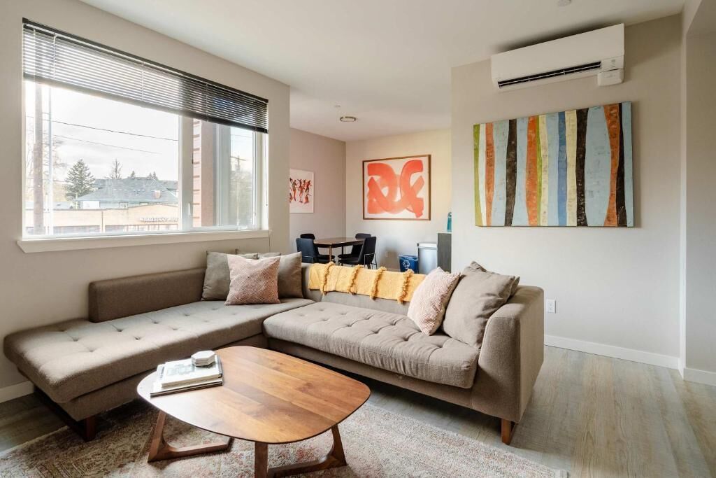 A living room with a sectional sofa, a coffee table, a window with blinds, an air conditioner, and artwork on the wall.