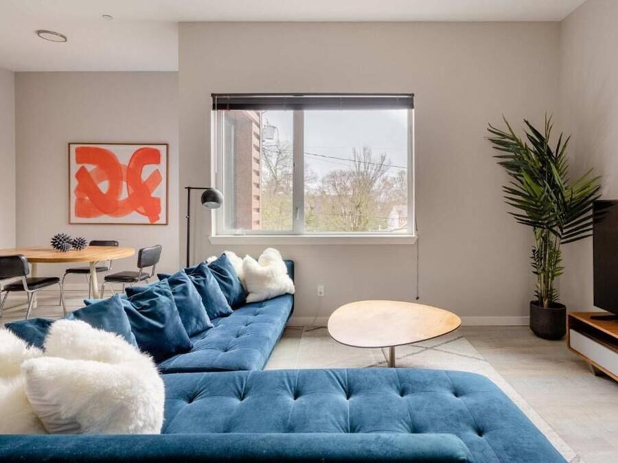 Ein modernes Wohnzimmer mit einem blauen Modulsofa, einem runden Couchtisch, einem Fernseher auf einem Holzständer, einem Essbereich mit Tisch und Stühlen sowie einem großen Fenster mit Blick auf Gebäude.