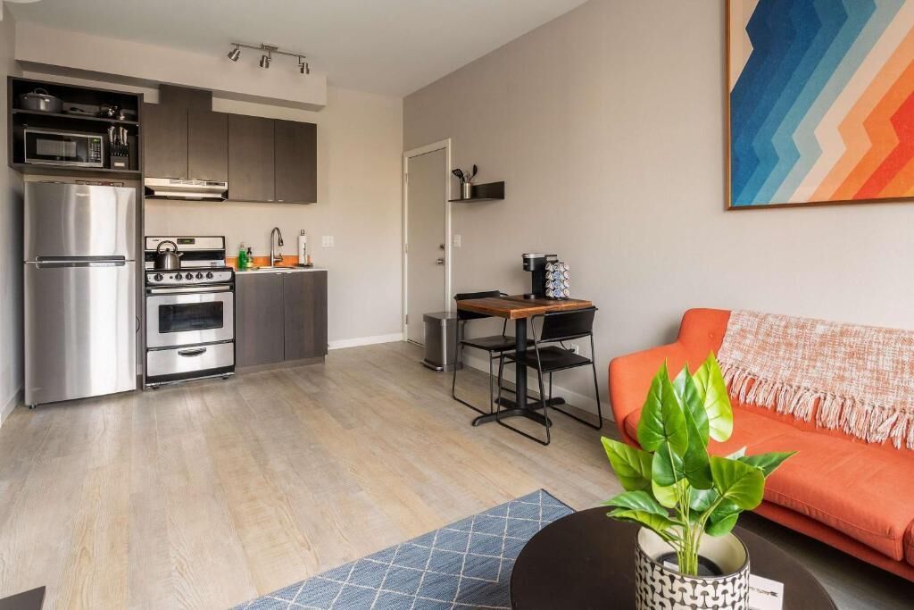 A modern kitchen with stainless steel appliances, a dining area with a table and chairs, and a living room with a sofa and a plant.