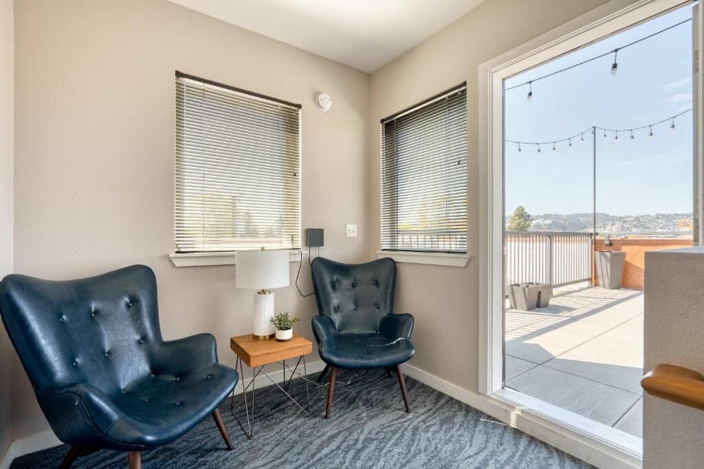 A room with two black leather chairs, a small table with a lamp, and a view through a large window to an outdoor balcony.
