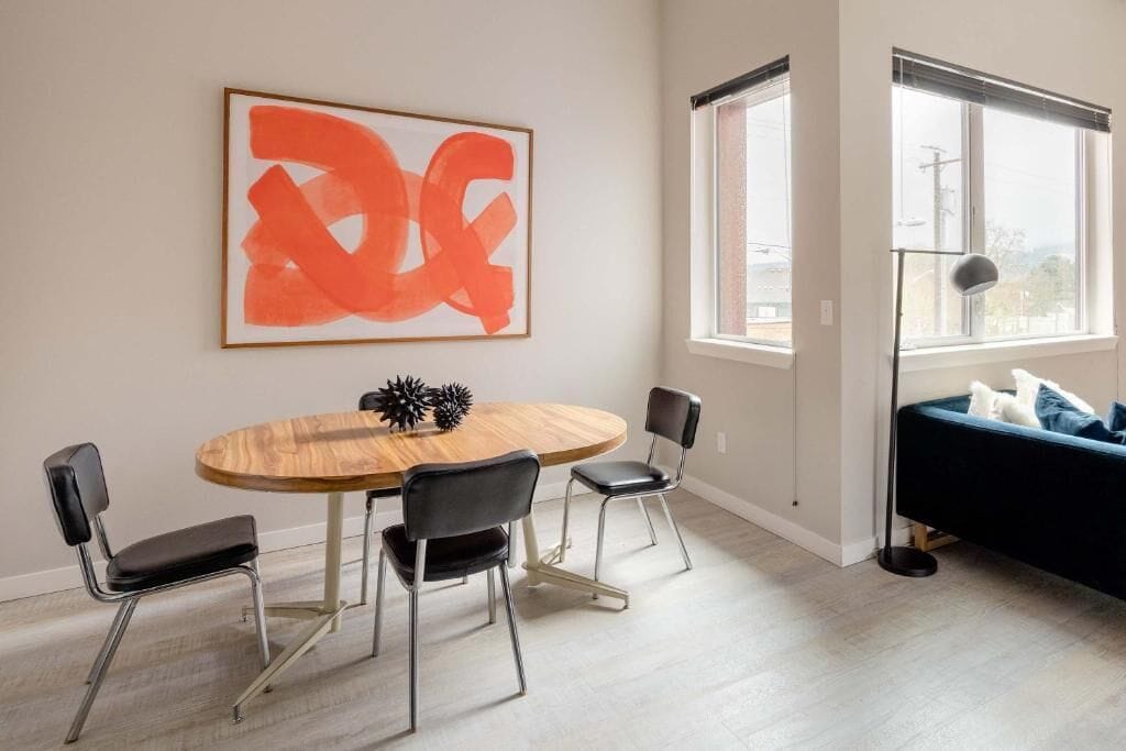 A modern living room with a round wooden table, black chairs, a blue sofa, a large abstract painting, and a window with blinds.