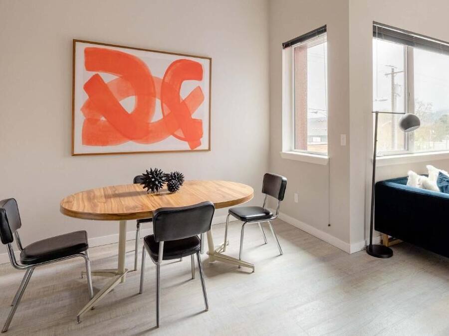 A modern living room with a round wooden table, black chairs, a blue sofa, a large abstract painting, and a window with blinds.