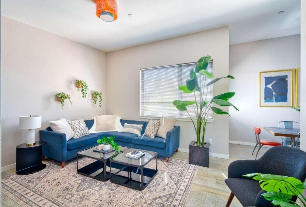 Una sala de estar con un sofá azul, una mesa de centro, una planta grande y un ventanal con persianas.