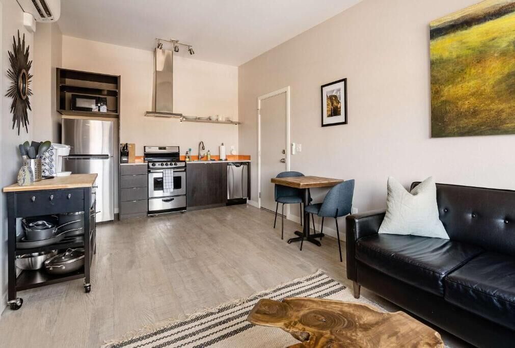A modern kitchen with a refrigerator, stove, and microwave, a dining area with a table and chairs, and a black leather sofa with a cushion.