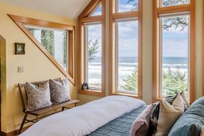 A bedroom with a large window, a bed, a bench, and a view of the ocean. - Private Oceanfront Luxury | Haystack Rock Views | Exclusive Chapman Point Retreat (Cannon Beach)