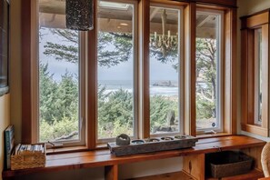 A room with large windows overlooking a forest and ocean, a wooden bench with a tray, and a chandelier. - Private Oceanfront Luxury | Haystack Rock Views | Exclusive Chapman Point Retreat (Cannon Beach)