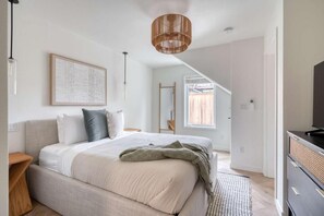 A bedroom with a bed, a bedside table, a hanging light fixture, a framed picture on the wall, and a window.