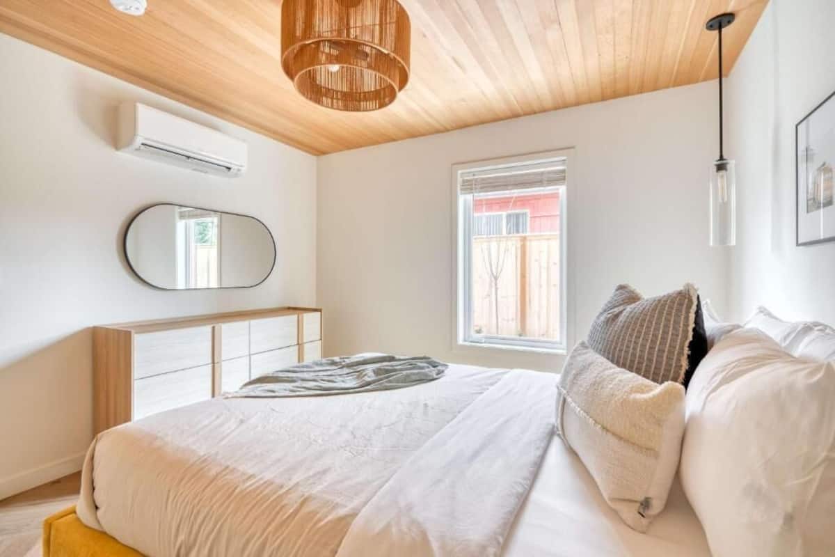 Un dormitorio con cama, espejo, equipo de aire acondicionado, ventana y lámpara colgante.