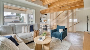 Een moderne woonkamer met een bank, fauteuil en een rond bijzettafeltje. Achtergrond toont een trap met houten leuningen.