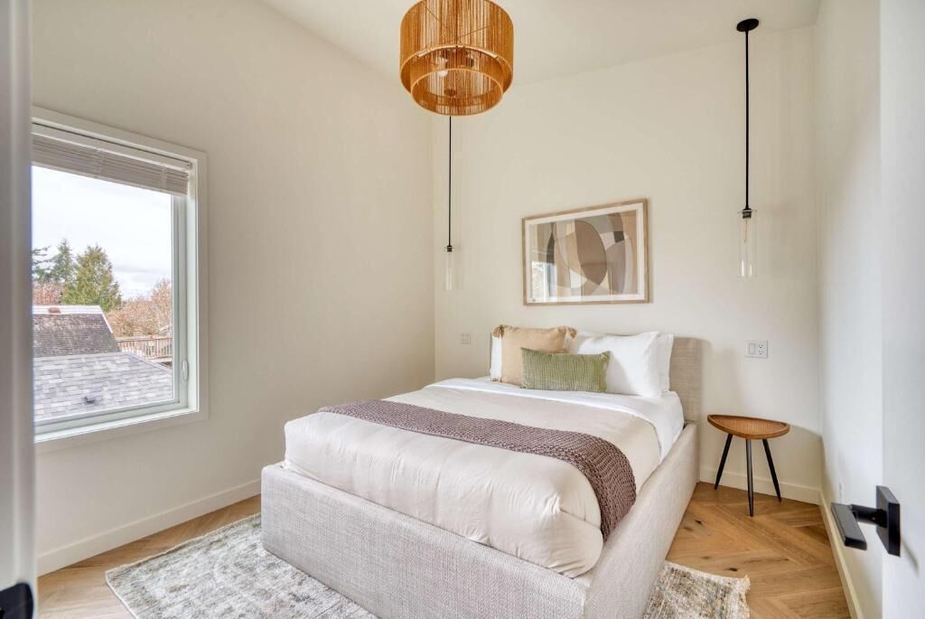 A bedroom with a large window, a bed with a headboard, a bedside table, and a hanging light fixture.