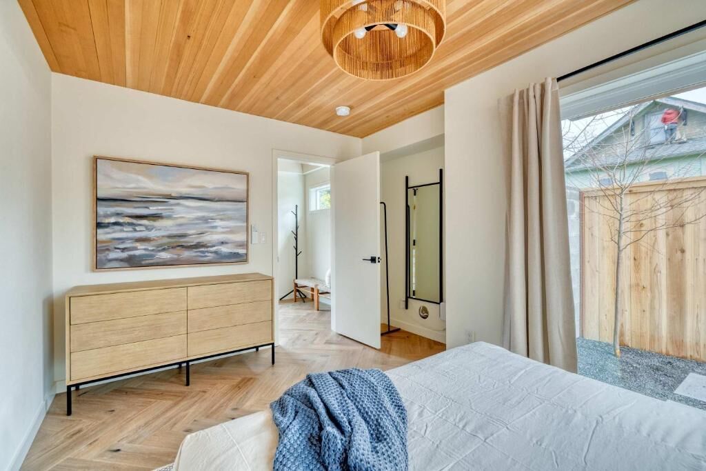 A bedroom with a bed, a wooden ceiling, a painting, a wardrobe, and a view of the outside.