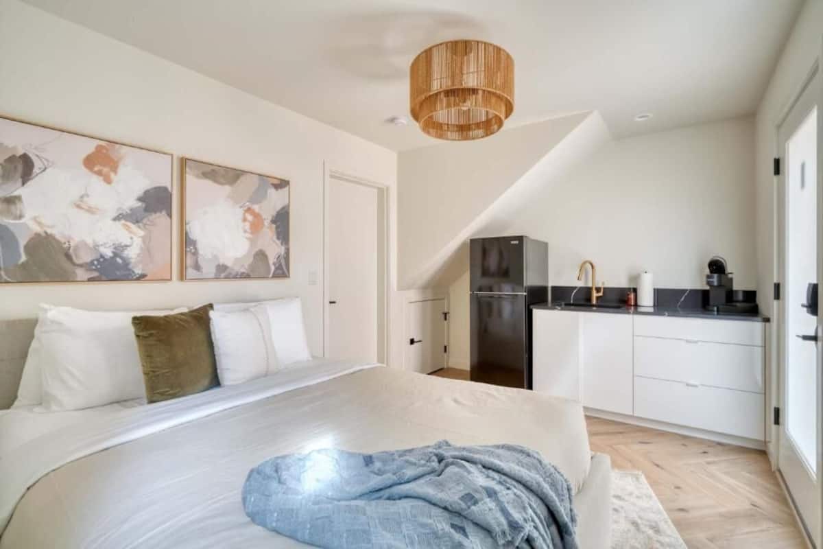 Un dormitorio con cama, un ventanal grande y una zona de cocina con armarios y electrodomésticos.