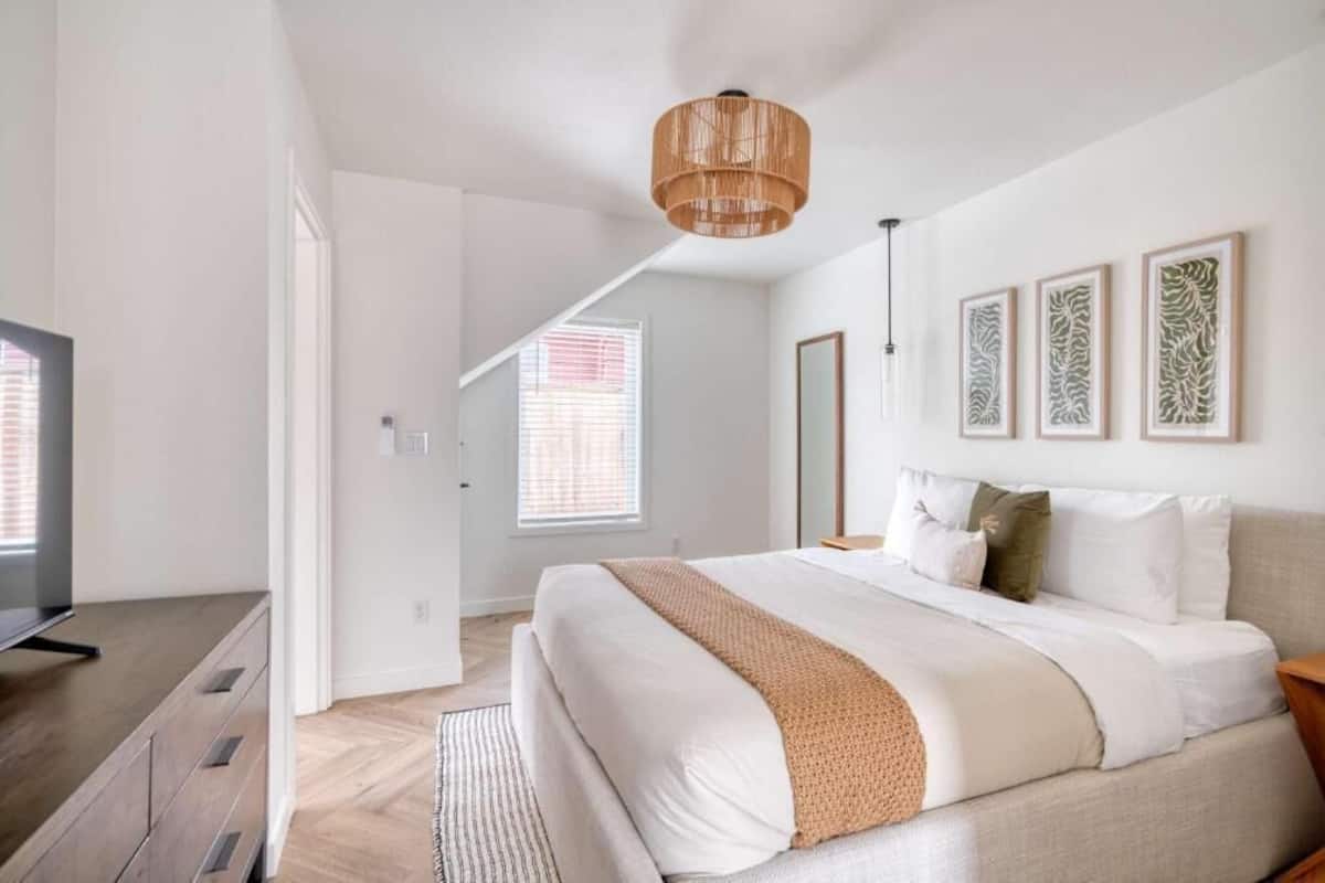 Un dormitorio con cama, televisión, suelo de madera, luz de techo y ventana con persianas.