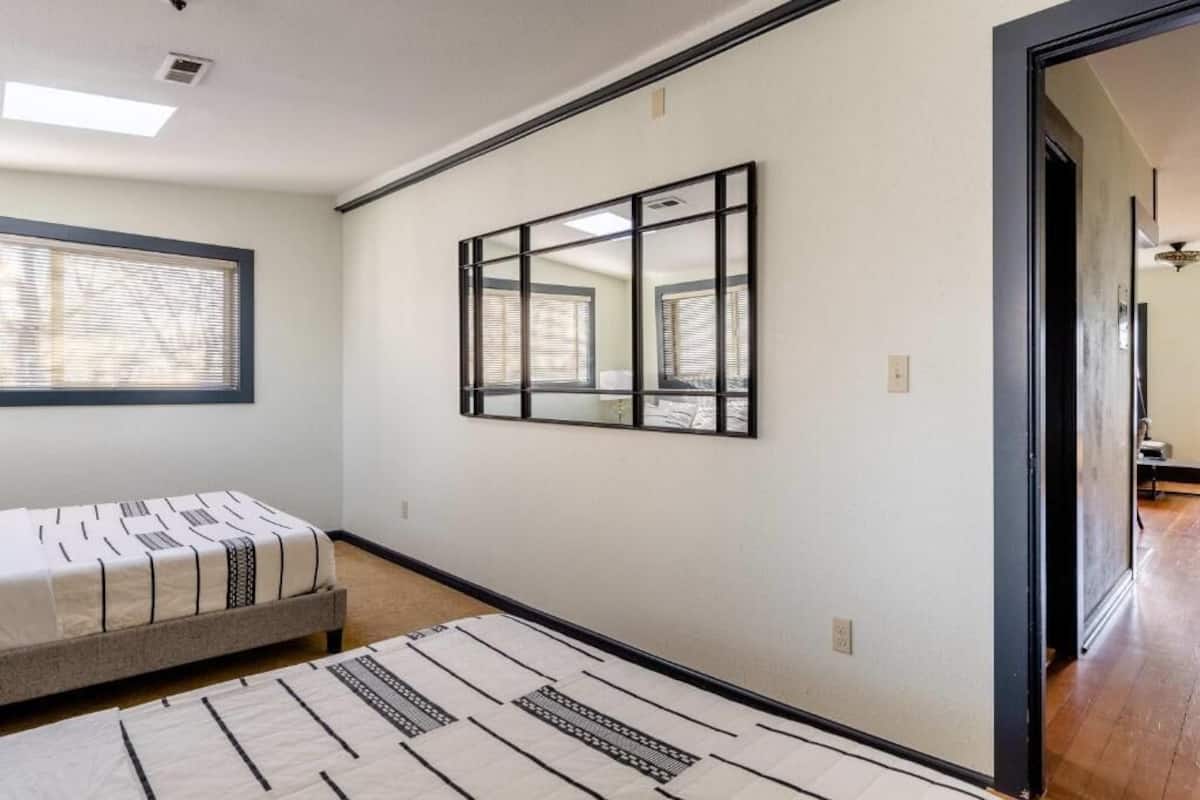 Un dormitorio con cama, ventana con persianas, espejo montado en la pared y puerta que da a otra habitación.
