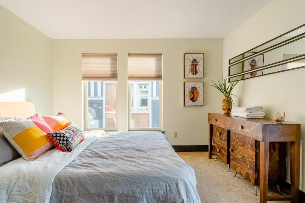 Une chambre à coucher comprenant un lit, une commode, une plante en pot et deux fenêtres équipées de stores.