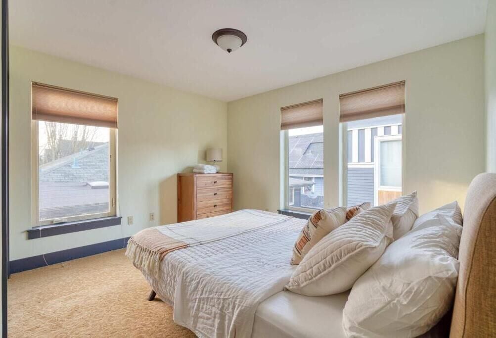 Une chambre à coucher avec un lit, deux fenêtres équipées de stores, une commode et un luminaire au plafond.