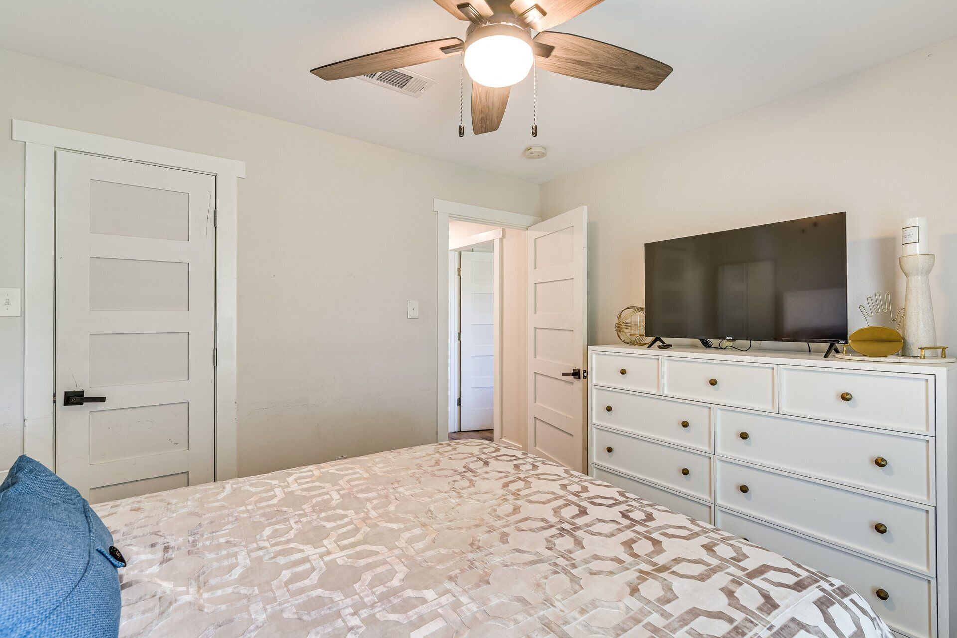 A bedroom with a bed, dresser, TV, ceiling fan, and a door leading to another room.