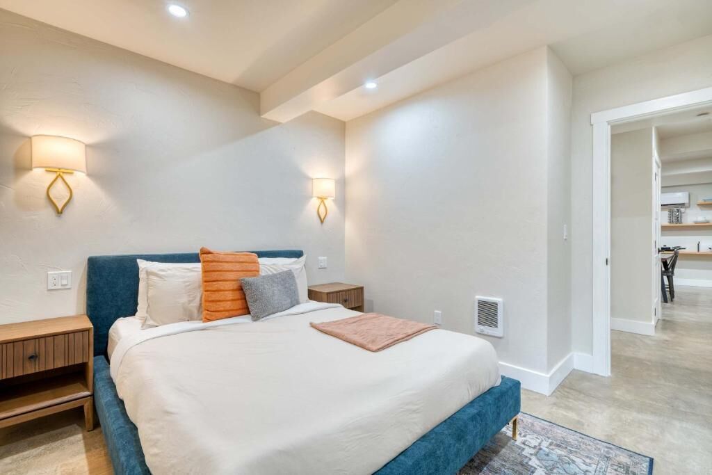 Une chambre à coucher comprenant un lit, des tables de chevet, des lampes fixées au mur et donnant sur la cuisine adjacente.