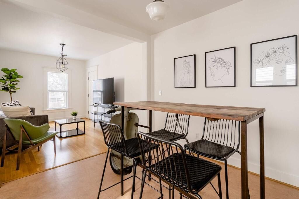 Un salon moderne avec une table à manger en bois, des chaises à structure métallique noire, un fauteuil vert, un téléviseur et des œuvres d’art encadrées accrochées au mur.
