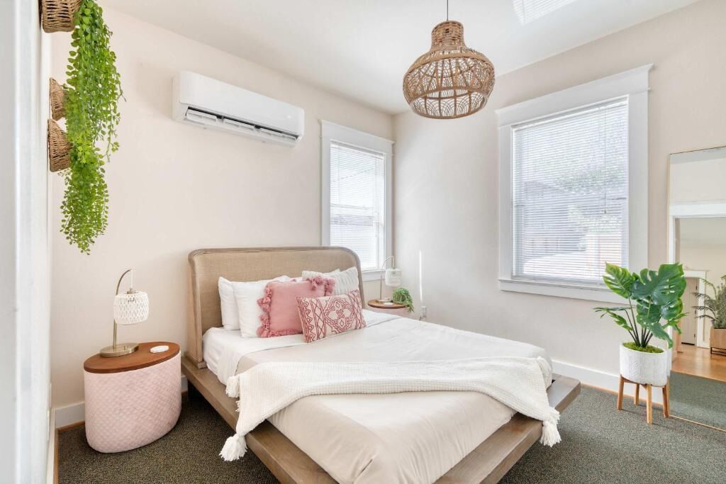 A bedroom with a bed, bedside tables, a hanging plant, an air conditioner, and a window.