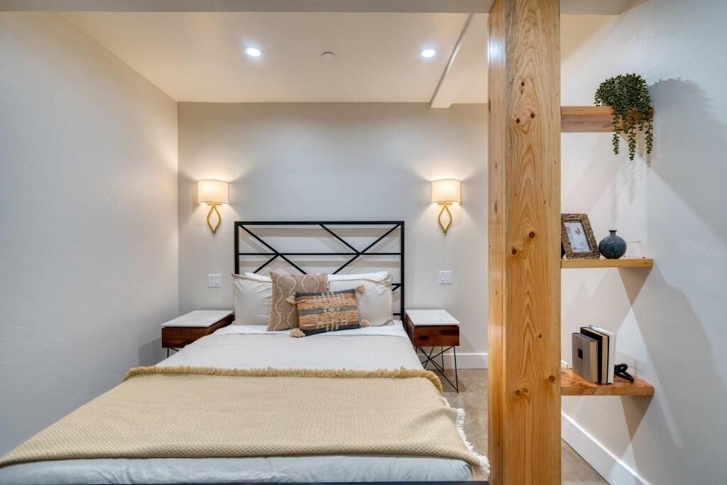 Un dormitorio con una cama, mesitas de noche, lámparas de pared, una viga de madera y un estante con libros y una planta.