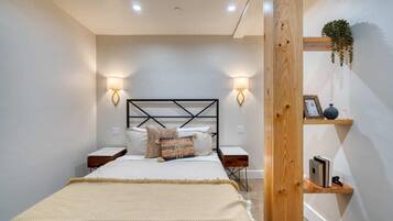 Un dormitorio con una cama, mesitas de noche, lámparas de pared, una viga de madera y un estante con libros y una planta.