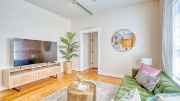 A living room with a flat-screen TV, wooden entertainment unit, green sofa, round coffee table, and a potted plant.