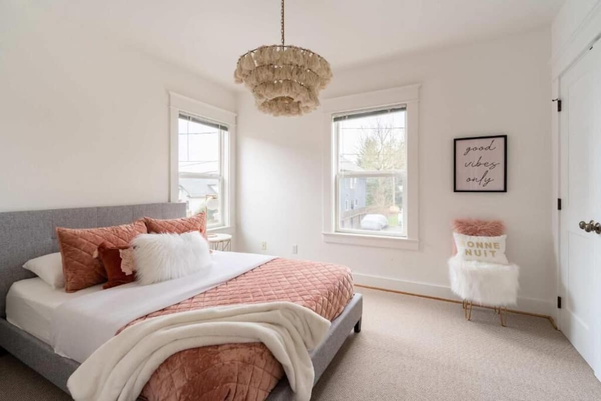 A bedroom with a bed, two windows, a chandelier, and a small stool with a cushion.