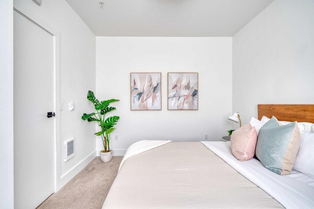 A bedroom with a bed, two wall-mounted paintings, a potted plant, and a bedside lamp.