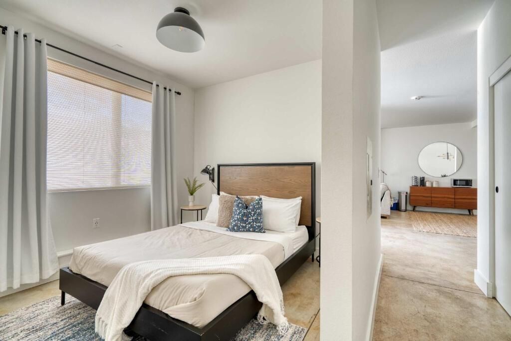 Une chambre à coucher moderne avec un grand lit, une table de chevet, une petite plante et une vue sur une autre pièce où se trouvent une commode en bois et un miroir rond accroché au mur.