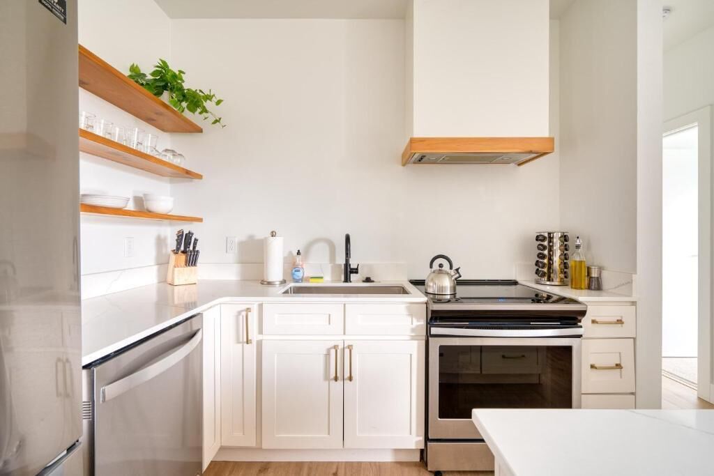 A modern kitchen with white cabinets, a stainless steel refrigerator, a built-in oven, and a wooden shelf with kitchen utensils.
