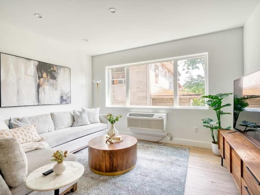 Ein modernes Wohnzimmer mit grauem Sofa, rundem Couchtisch, Flachbildfernseher und großem Fenster mit Jalousien.