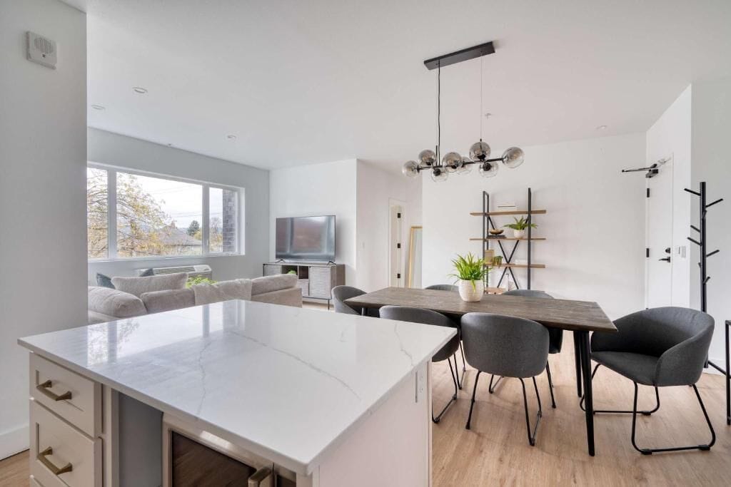 A modern kitchen with a large island, dining table, and chairs, a flat-screen TV, and a window with a view.