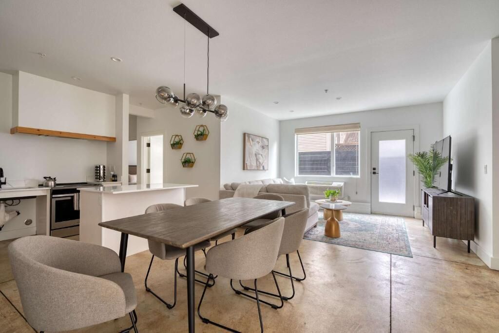 A modern kitchen with a dining area, a living room with a sofa and TV, and a large window.