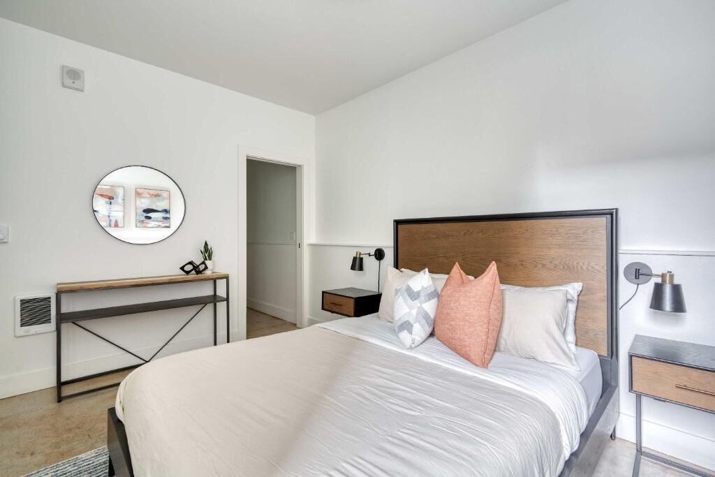 A modern bedroom with a large bed, a bedside table, a round mirror, and a small desk.