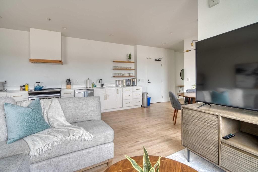 A modern living room with a sofa, a coffee table, a TV, and a kitchen in the background.
