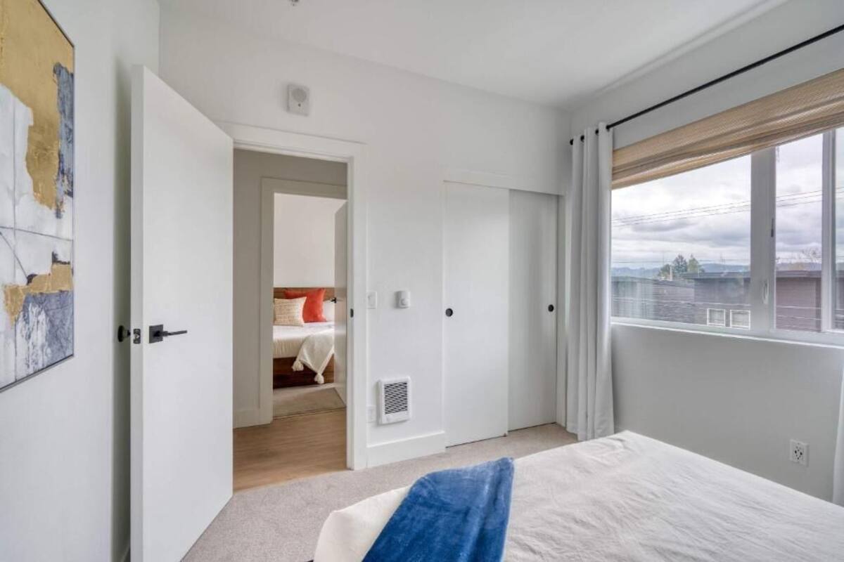 A bedroom with a bed, a window with curtains, a door leading to another room, and a wall with a painting.