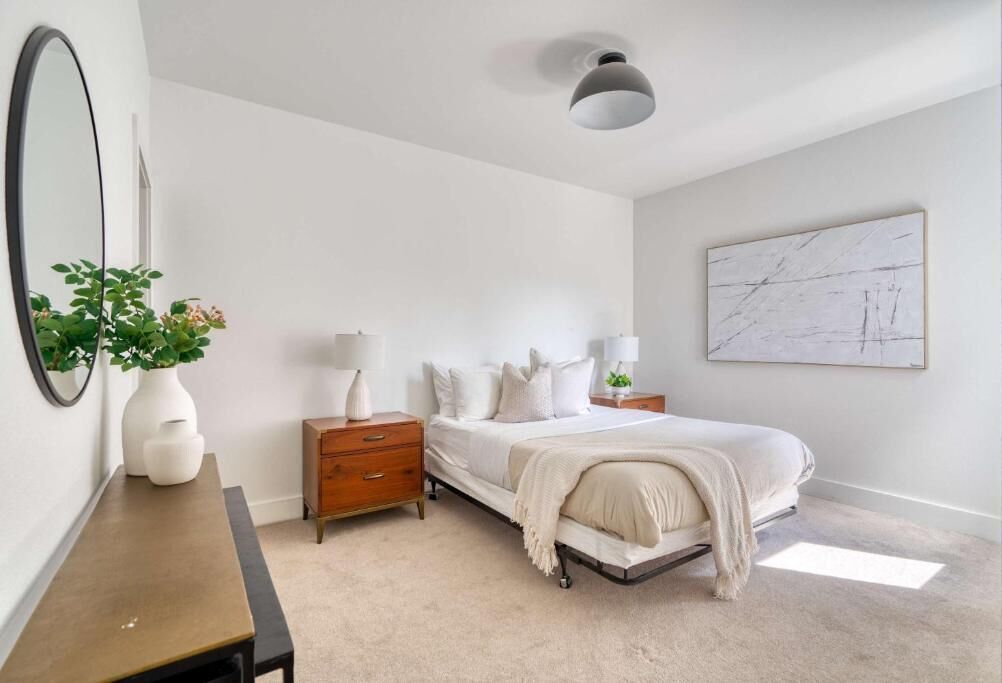 A bedroom with a bed, bedside tables, a dresser, a mirror, a plant, and a ceiling light fixture.