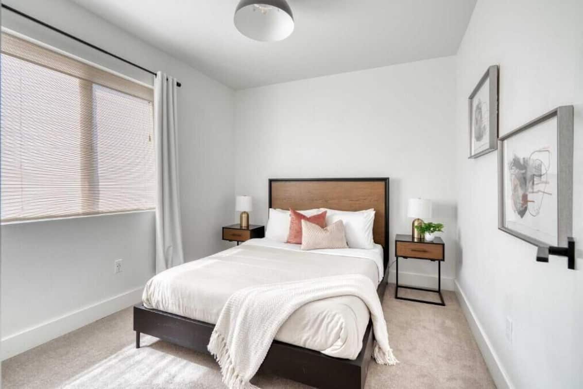 A bedroom with a bed, bedside tables, a nightstand, a window with blinds, and wall-mounted artwork.