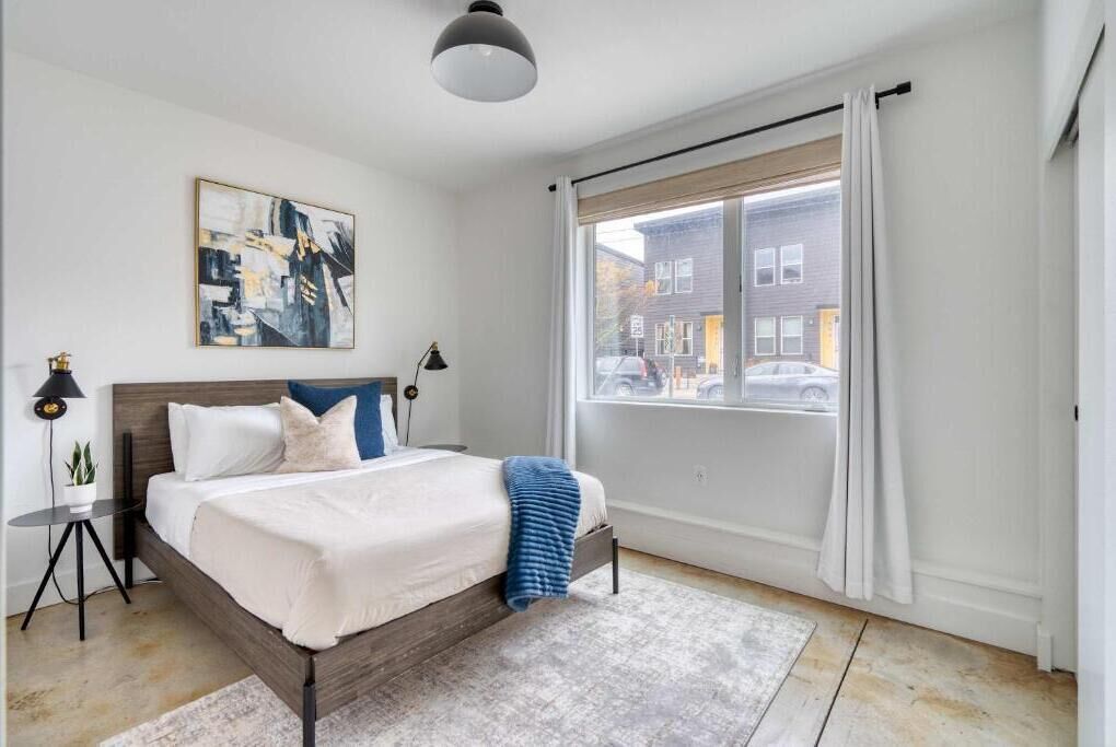 A bedroom with a bed, a window with curtains, a painting on the wall, and a small table with a lamp.