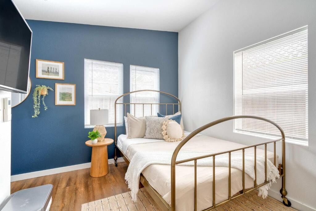 Un dormitorio con cama, televisión, dos ventanas, una mesita con una planta y una pared con cuadros.