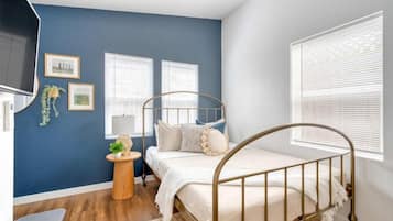 Un dormitorio con cama, televisión, dos ventanas, una mesita con una planta y una pared con cuadros.
