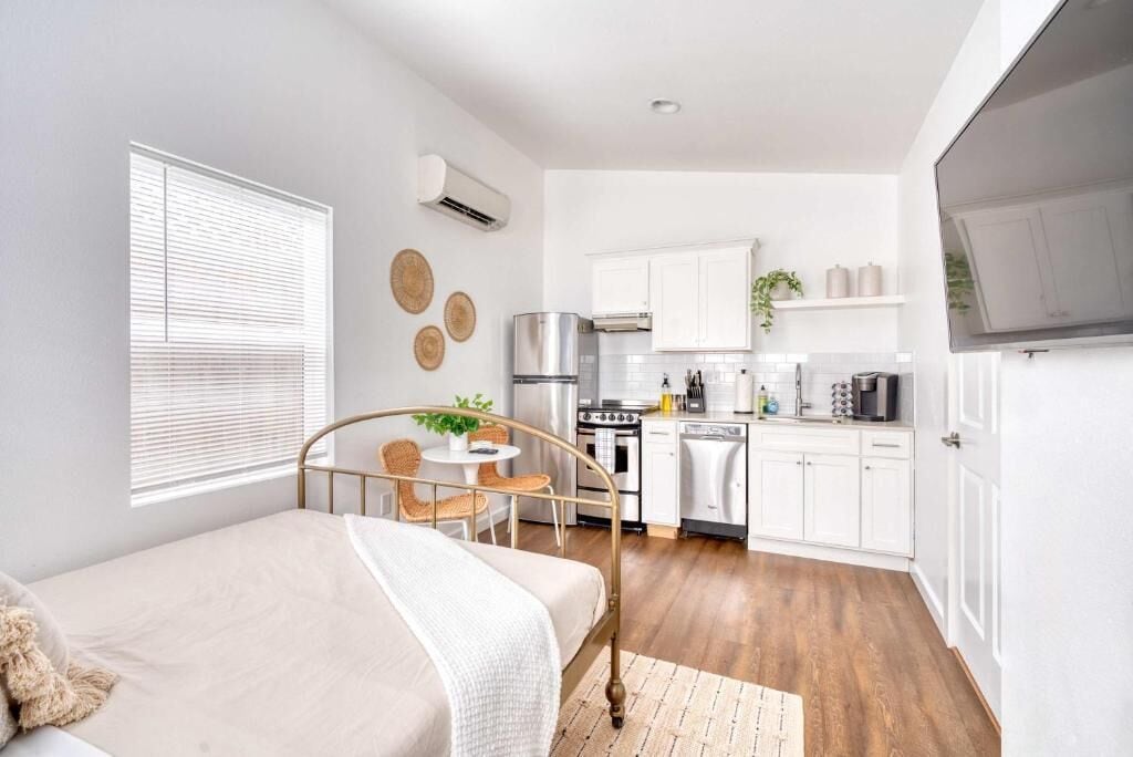 A compact bedroom with a bed, bedside table, and a small seating area. A kitchenette with a stove, microwave, and cabinets is visible.